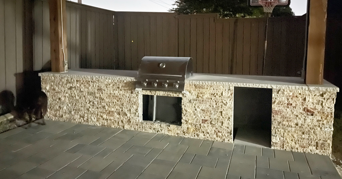 Picture of outdoor kitchen. Progress shows cinderblocks with outer layer of stonework application & gas grill.
